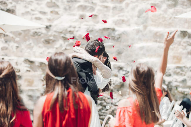 Молодой жених и невеста целуются на свадебной церемонии под лепестками роз, брошенными гостями — стоковое фото