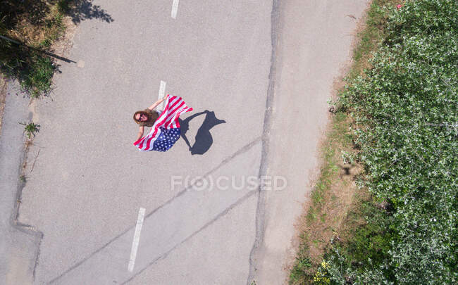Mulher de chapéu a andar com a bandeira americana numa estrada solitária. Dia especial para comemorar o 4 de julho — Fotografia de Stock