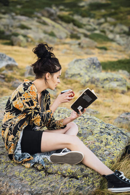 Вид сбоку на молодую женщину за чашкой кофе, сидящую на камнях в ванне и читающую книгу — стоковое фото