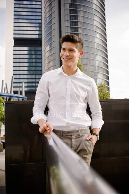 Sonriente joven hombre de negocios en traje elegante sonriendo y mirando hacia otro lado mientras está de pie cerca de barandilla en la calle de la ciudad moderna - foto de stock