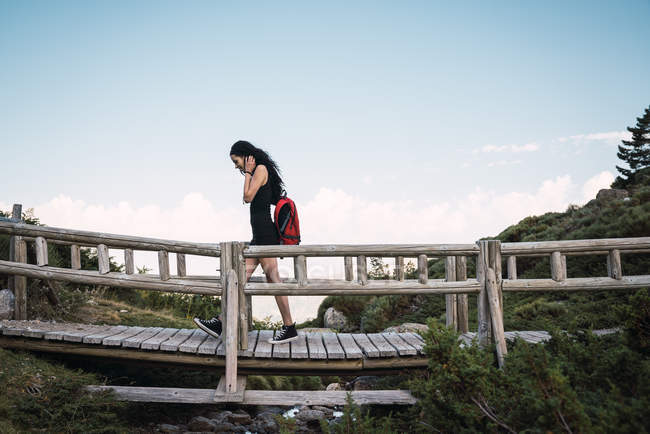 Вид сбоку случайной женщины с красным рюкзаком, идущей по деревянному пешеходному мосту в зеленом против голубого неба — стоковое фото