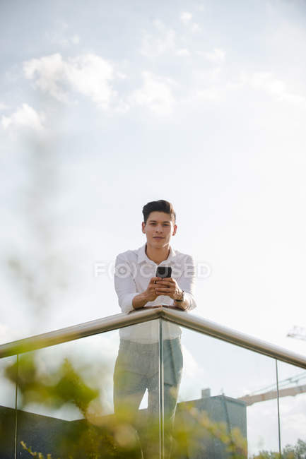 Young guy in casual outfit holding smartphone and looking at camera while leaning on railing on background of cloudy sky — Stock Photo