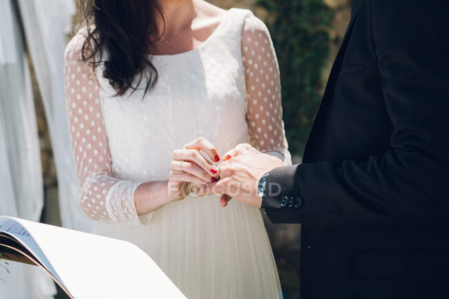 Вид на урожай молодой невесты в свадебном платье и жениха в черном костюме, обменивающихся кольцами на церемонии в саду — стоковое фото