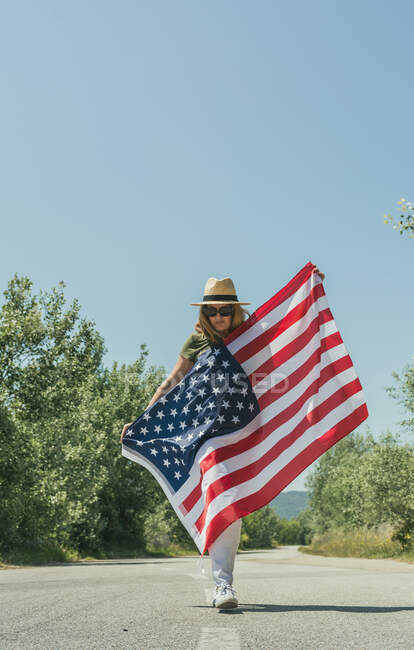 Mulher feliz andando com uma bandeira americana e celebrando em uma estrada solitária. Dia especial para comemorar o 4 de julho — Fotografia de Stock