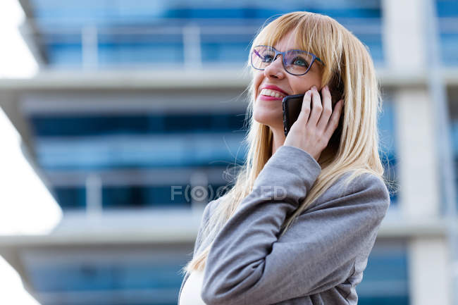 Mujer rubia en chaqueta gris y gafas hablando por teléfono móvil con centro de negocios en el fondo - foto de stock