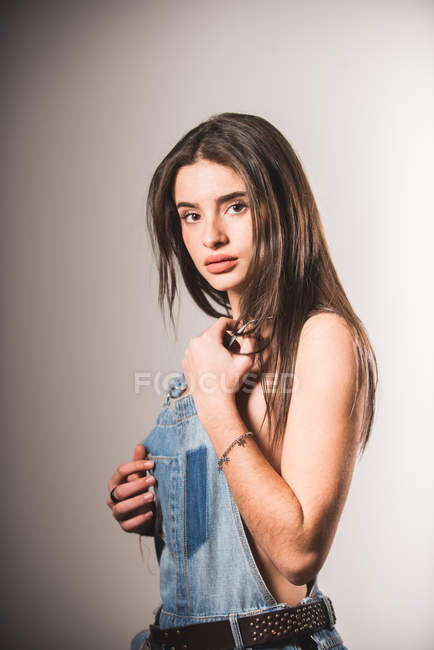 Menina sexy em macacão jeans sobre corpo nu posando no fundo cinza — Fotografia de Stock