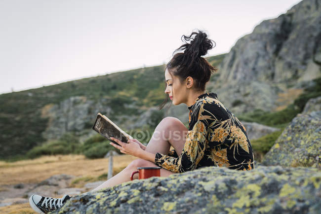 Young brunette woman reading book on stones while traveling — Stock Photo