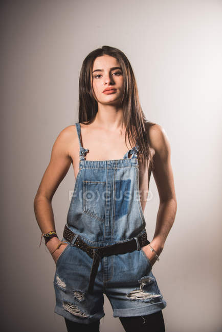 Сексуальная девушка в джинсовом комбинезоне над обнаженным телом позирует на сером фоне — стоковое фото