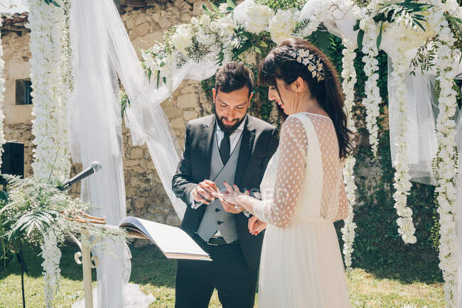 Joven novia hermosa en vestido blanco y novio guapo en traje negro intercambio de anillos en el fondo de las decoraciones de la boda - foto de stock