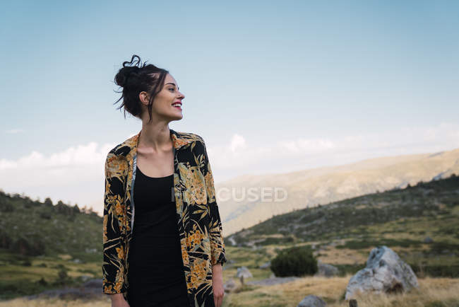 Hermosa morena en camisa colorida sonriendo mientras está de pie en el verde valle rocoso de las montañas, España - foto de stock