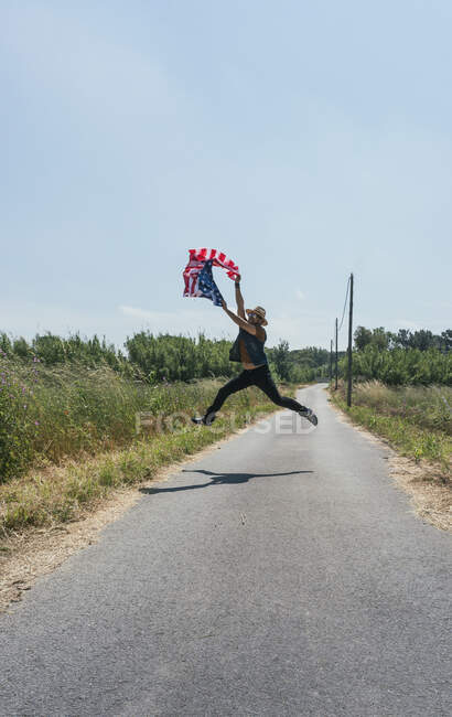 Uomo felice e che salta per la gioia con una bandiera americana su una strada solitaria — Foto stock