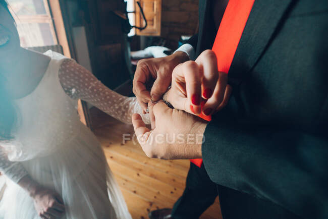 Crop Ansicht des Mannes in schwarzem Kostüm hilft junge Braut im Brautkleid auf Armband auf der Hand auf dem Hintergrund des Zimmers setzen — Stockfoto