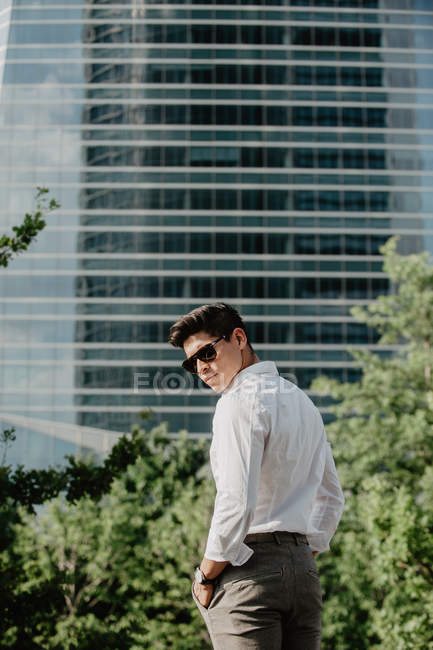 Молодий бізнесмен в сонцезахисних окулярах стоїть перед сучасною будівлею і дивиться через плече — стокове фото