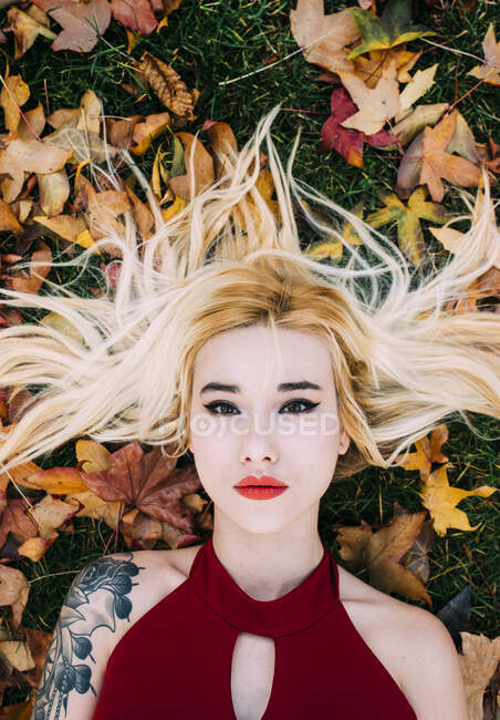 From above blonde woman with tattooed arm lying on green grass in foliage and looking at camera. — Stock Photo