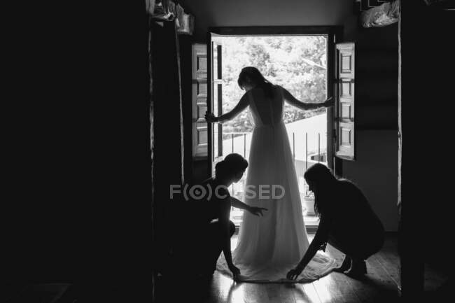 Back view of bride standing near window and two women helping with floor length white wedding dress in black and white colors - foto de stock