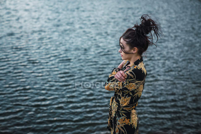 Vista lateral de la mujer en camisa colorida abrazándose y mirando hacia otro lado de pie sobre un fondo borroso de agua azul en ondas - foto de stock