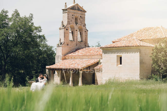 Young man in black costume holding young woman in wedding dress in arms on background of green trees and old stone church at sunny day — Stock Photo