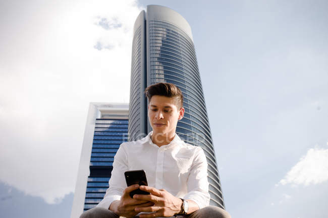 Молодий хлопець у повсякденному вбранні, використовуючи смартфон на тлі хмарного неба та сучасного хмарочоса — стокове фото