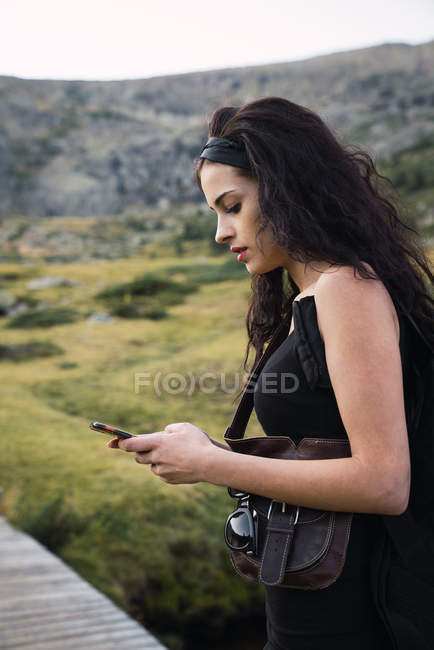 Side view of beautiful brunette with backpack standing on walkway in green mountains and using phone — Stock Photo