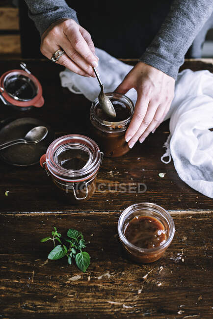 From above shot of hands of anonymous woman using spoon to take thick sauce from jar while standing near wooden table — Stock Photo