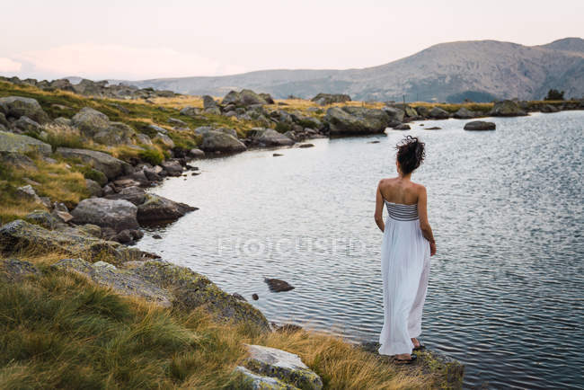 Young woman in white dress standing alone on shore of lake — Stock Photo