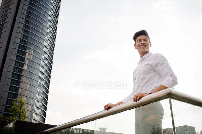 Усміхнений молодий бізнесмен в елегантному вбранні посміхається і дивиться геть, стоячи біля перил на вулиці сучасного міста — стокове фото
