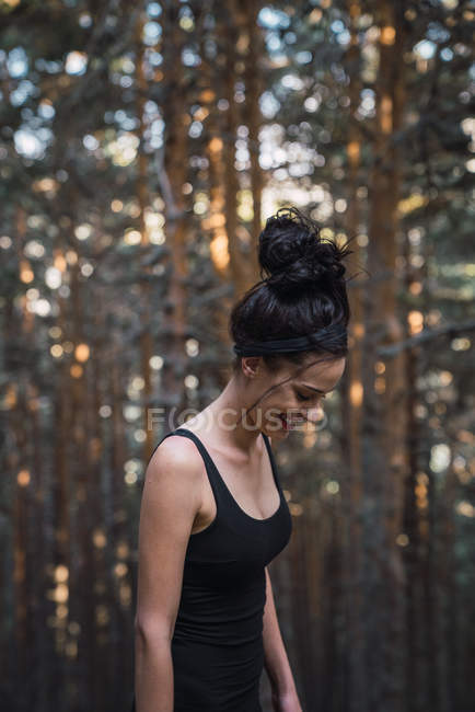 Beautiful young woman in tank top laughing and looking down while standing in woods — Stock Photo