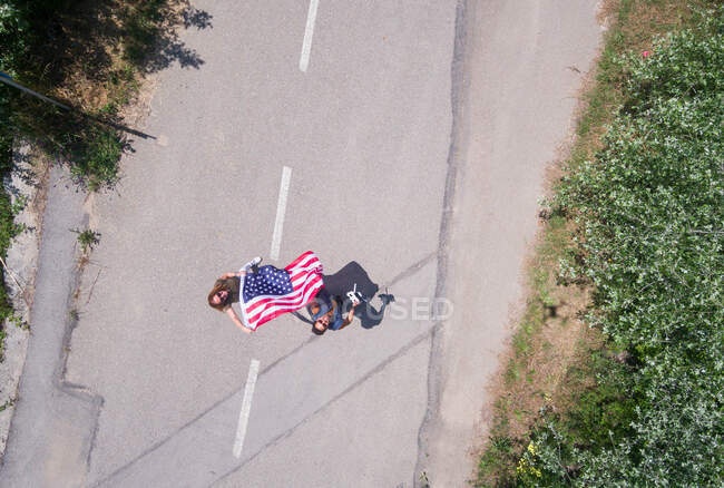 Femme au chapeau marchant avec le drapeau américain sur une route solitaire. Journée spéciale pour célébrer le 4 juillet — Photo de stock