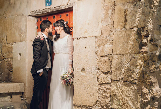 Heureuse belle mariée en robe de mariée tenant bouquet et marié en costume noir souriant et s'embrassant près du mur de pierre — Photo de stock