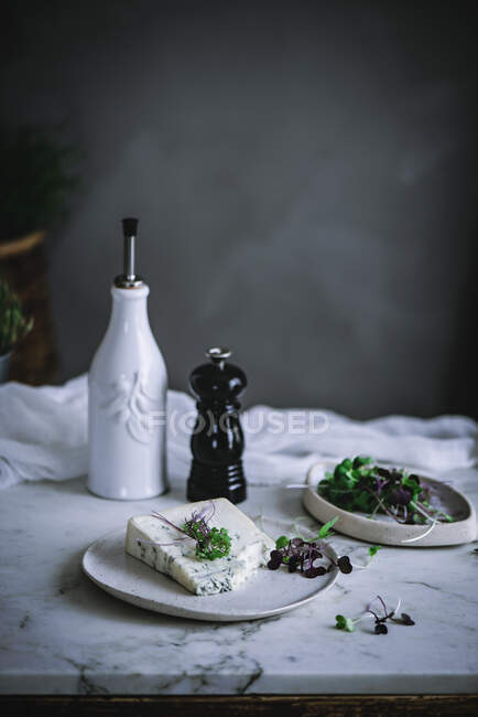 Blue cheese with herbs in white dish standing on marble table designed with vessels and white textile material in soft focus — Stock Photo