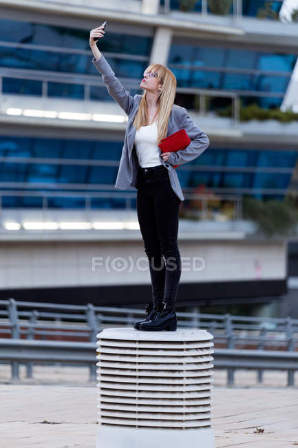 Блондинка в серой куртке и черных джинсах делает селфи с телефоном на улице — стоковое фото