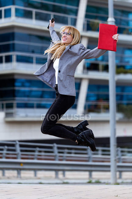 Blond woman in glasses and gray jacket with red bag and phone jumping in front of modern building — Stock Photo