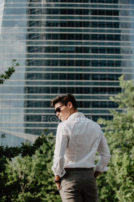 Молодой бизнесмен в солнечных очках, стоящий перед современным зданием и оглядывающийся через плечо — стоковое фото