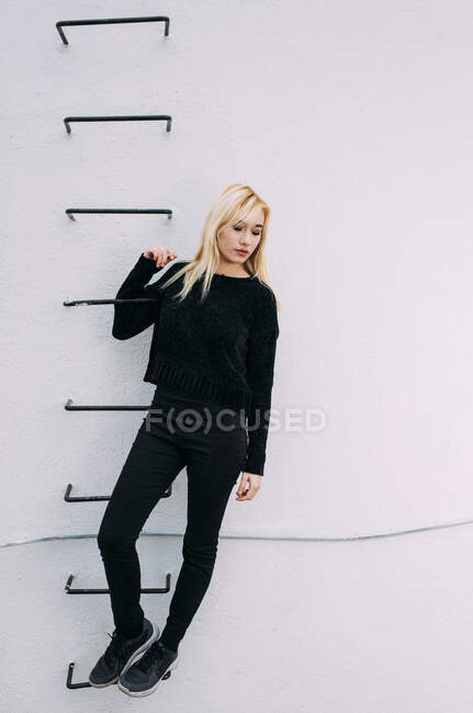 Junge hübsche blonde Frau hält sich auf Leiter an der weißen Wand. — Stockfoto