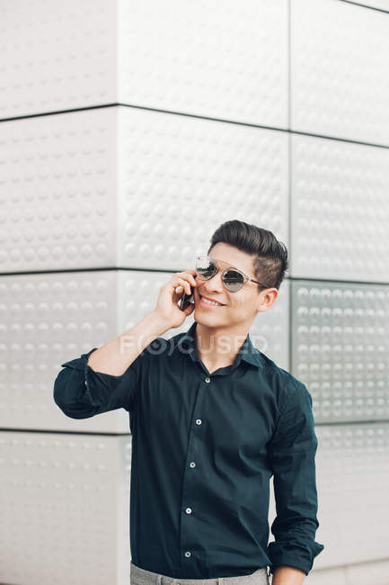 Joven empresario alegre hablando por teléfono contra la pared de la construcción - foto de stock