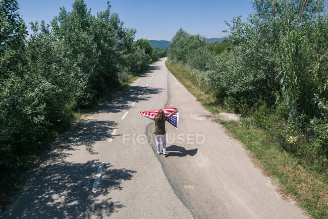 Mulher de chapéu a andar com a bandeira americana numa estrada solitária. Dia especial para comemorar o 4 de julho — Fotografia de Stock