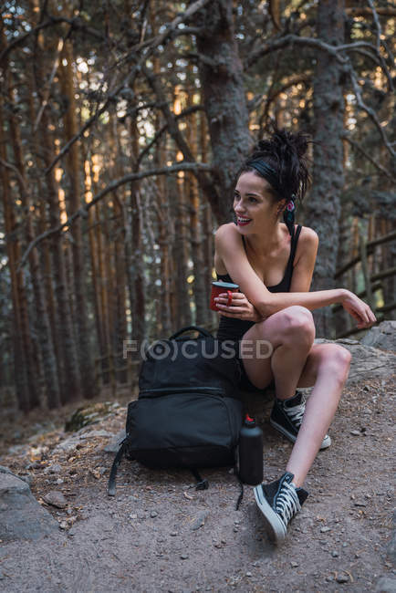 Очаровательная женщина с рюкзаком и чашей отдыхает в лесу в горах — стоковое фото