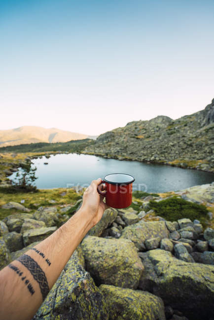 Cropped of tattooed hand of man with mug on shore of lake — Stock Photo