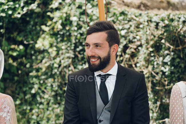 Joven novio guapo con cabello oscuro y barba sentado afuera y sonriendo en el fondo de arbustos verdes - foto de stock
