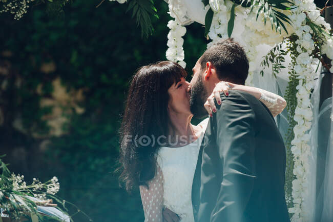 Joven novio guapo y hermosa novia besándose en la ceremonia de boda en el fondo de los árboles y decoraciones - foto de stock