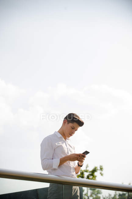 Joven empresario seguro de sí mismo usando el teléfono inteligente de pie en la barandilla - foto de stock