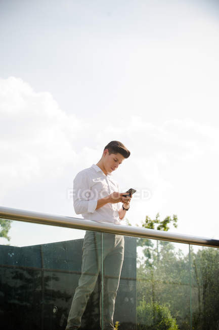Впевнений молодий бізнесмен за допомогою смартфона, що стоїть на перилах — стокове фото