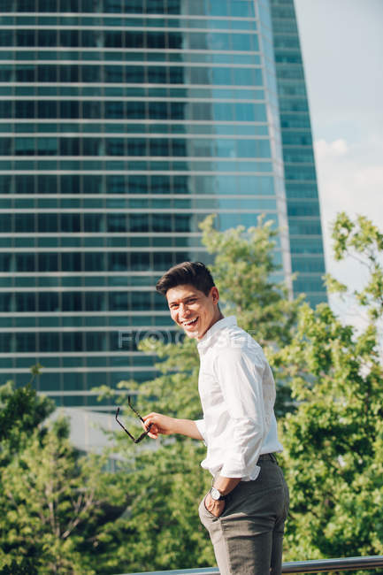 Rindo jovem empresário de pé contra edifício moderno e olhando sobre o ombro — Fotografia de Stock