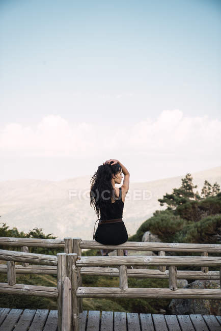 Rückansicht einer sinnlichen Frau in Schwarz mit langen Haaren, die auf einem Holzzaun über der malerischen Tallandschaft sitzt — Stockfoto