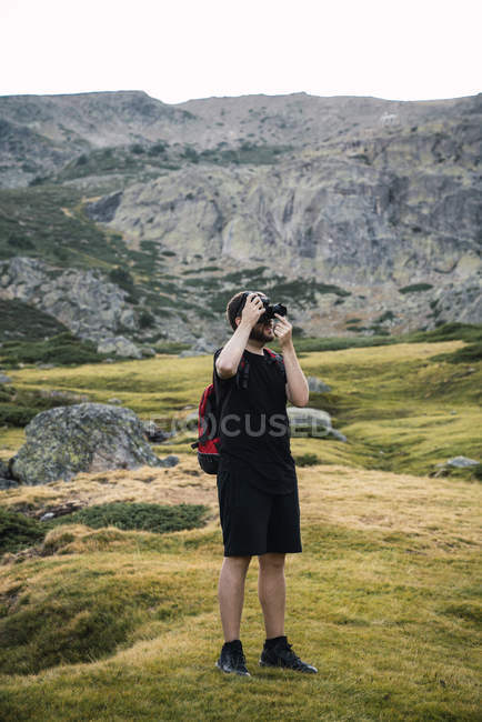 Homme adulte en tenue décontractée noire portant un sac à dos et prenant des photos de montagnes — Photo de stock