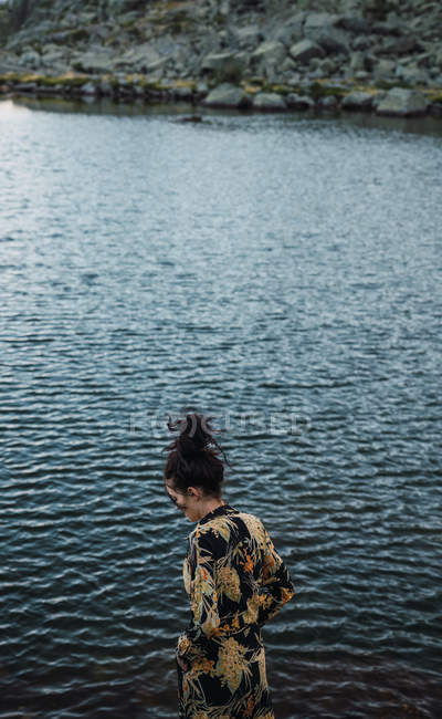 Young woman in colorful shirt standing alone on shore of lake — Stock Photo