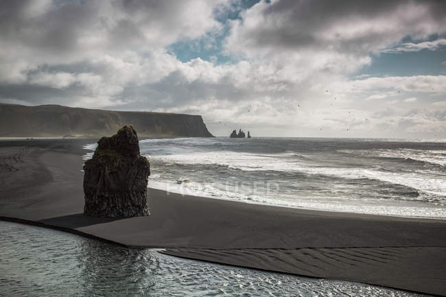 Roca en la playa con acantilados bajo el cielo dramático, Islandia - foto de stock