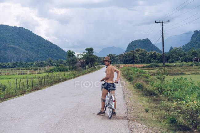 Shirtless man sitting on bicycle and looking back on rural road in hillside. — Stock Photo