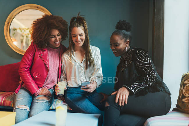 Grupo de tres hermosas mujeres jóvenes multirraciales sentadas en una cafetería interactuando en el teléfono móvil - foto de stock