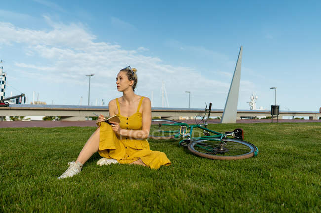 Mulher elegante em vestido amarelo fluindo e tênis sentado no gramado com bicicleta e pequeno caderno — Fotografia de Stock